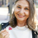 Close up outdoor picture of gorgeous positive trendy looking retired woman in her sixties having walking in park, holding ice cream cone and stick, offering you to have a ittle bit. Vertical image